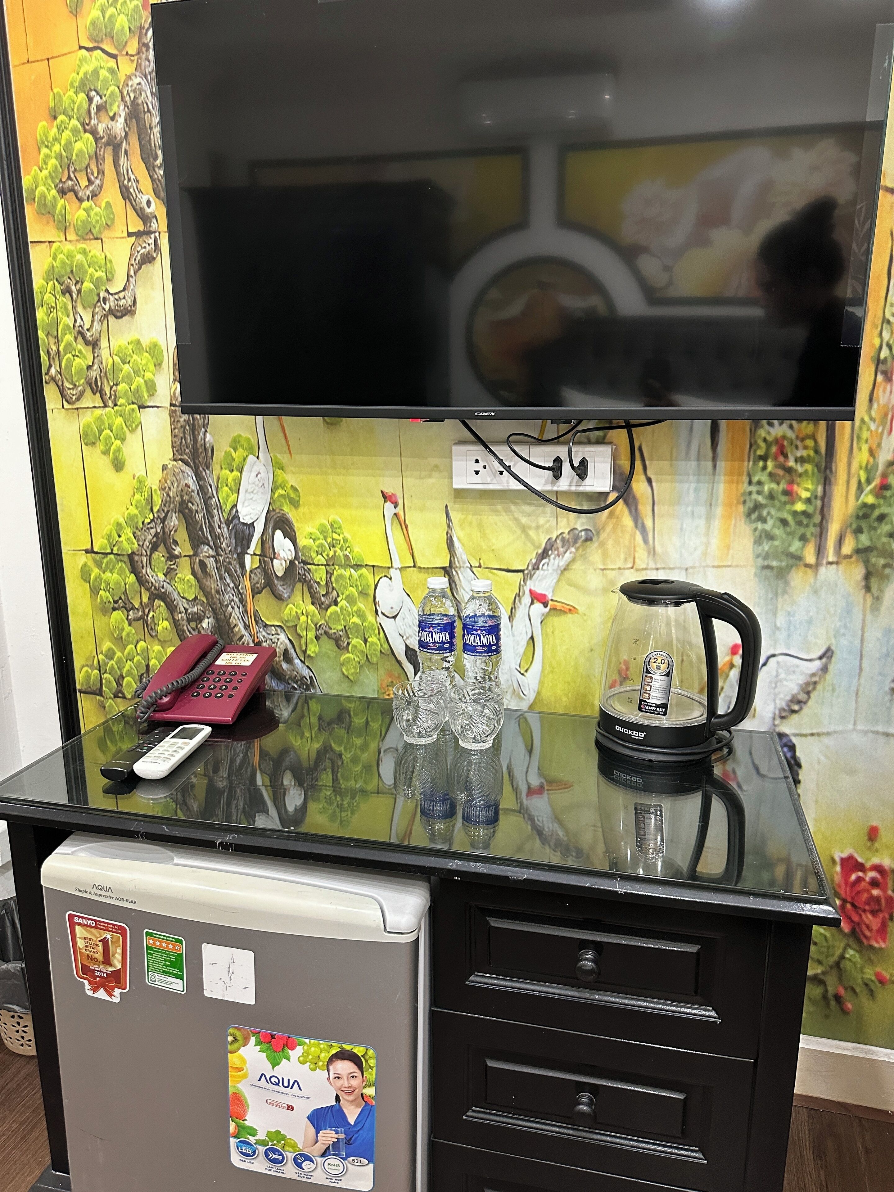 A room with a glass-top desk, a telephone, a water dispenser, and a television mounted above the desk.