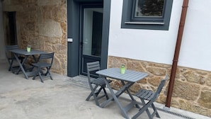 Un espacio al aire libre con dos mesas y cuatro sillas, un muro de piedra y una puerta con un ventanal encima.