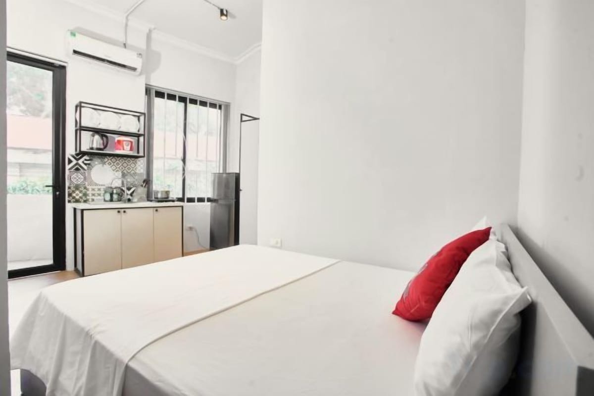 Una habitación con una cama, un almohadón rojo, un almohadón blanco, una zona de cocina con armarios y un refrigerador, y una ventana.