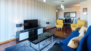 A modern living room with a flat-screen TV, a glass coffee table, a sofa, and yellow armchairs. There is a dining area with a table and chairs in the background. - GuestReady - Grandiose Getaway in Caniço (Caniço)