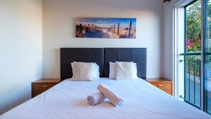 A neatly made bed with white linens and two pillows, a painting of a beach scene above it, and a window with a view of greenery and a balcony railing. - GuestReady - Grandiose Getaway in Caniço (Caniço)