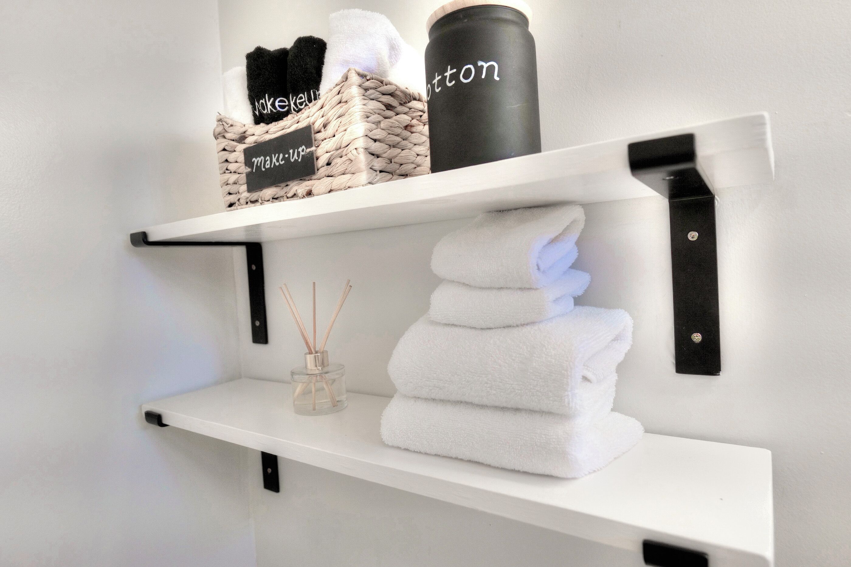 Een plank met een gevlochten mandje met de tekst ‘make-up’, een zwarte container met ‘cotton’ erop, en een stapel witte handdoeken.