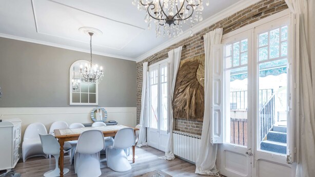 A dining area with a wooden table and white chairs, a chandelier, and a view of the outside through large windows. - Eixample 3BD Apartment with Private Terrace (Barcelona)