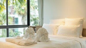 A neatly made bed with white linens and towels, a window with a view of palm trees and buildings, and a bedside table with a lamp. - 100mBeachfront gym Swimming Pools c100 (Rawai)