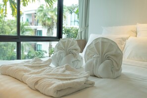 A neatly made bed with a white blanket and two rolled towels, a window with a view of greenery and buildings, and a white pillow on the bed.