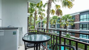 A balcony with a glass table and chairs, overlooking a swimming pool and palm trees, with a modern building in the background. - 100mBeachfront gym Swimming Pools c100 (Rawai)