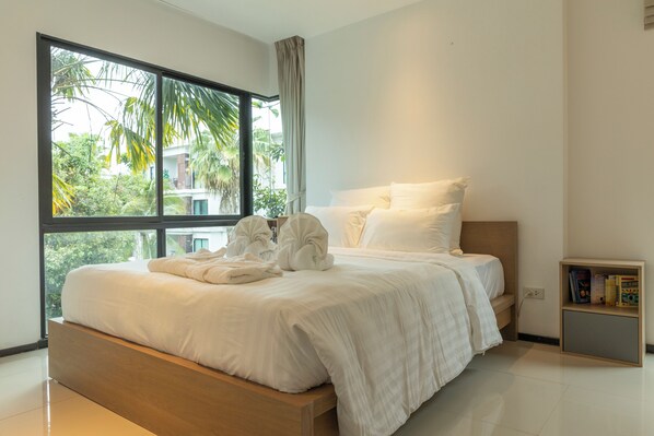 A modern bedroom with a large window, a wooden bed, white bedding, and a bedside table with books.