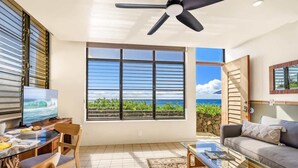 A modern living room with a large window offering a view of the ocean, a ceiling fan, and a dining area with a table and chairs. - Poipu Shores 101B - Ocean Front, 1BR, 1.5 bath (Koloa)