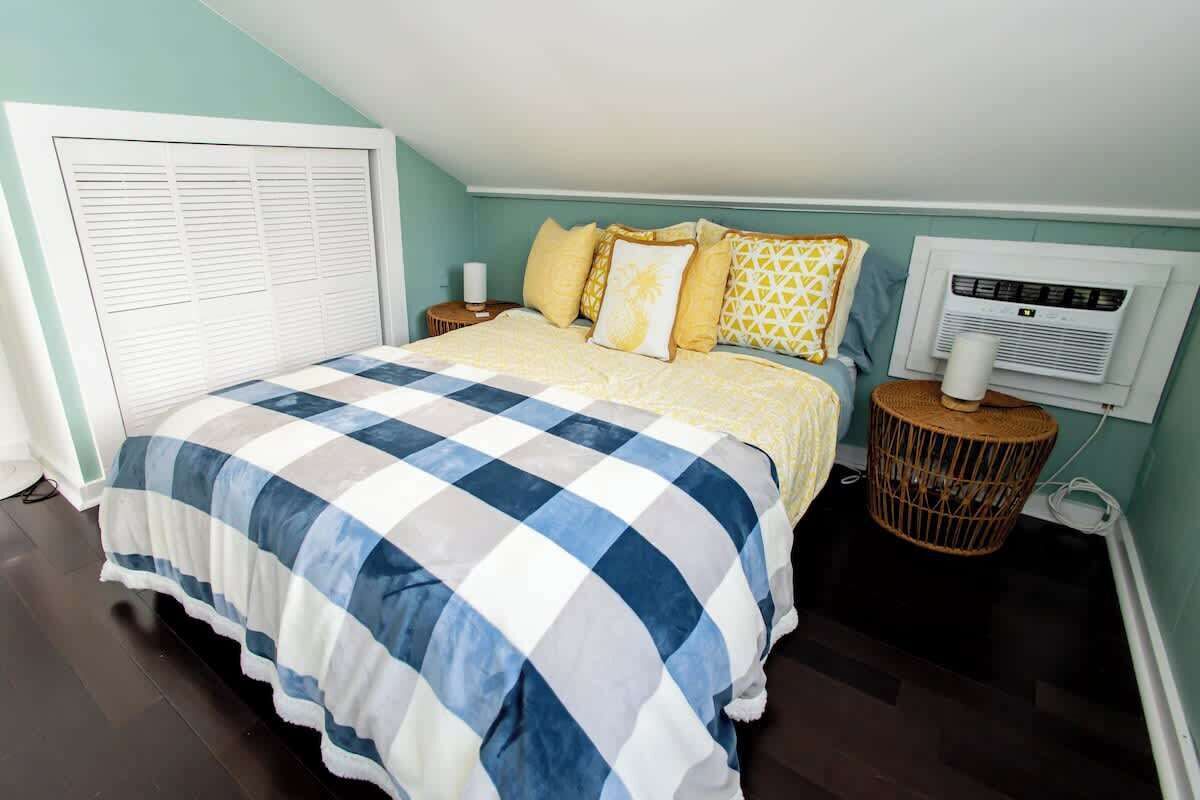 A bedroom with a checkered bedspread, a bedside lamp, a wall-mounted air conditioner, and a wicker nightstand.