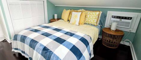 A bedroom with a checkered bedspread, a bedside lamp, a wall-mounted air conditioner, and a wicker nightstand.