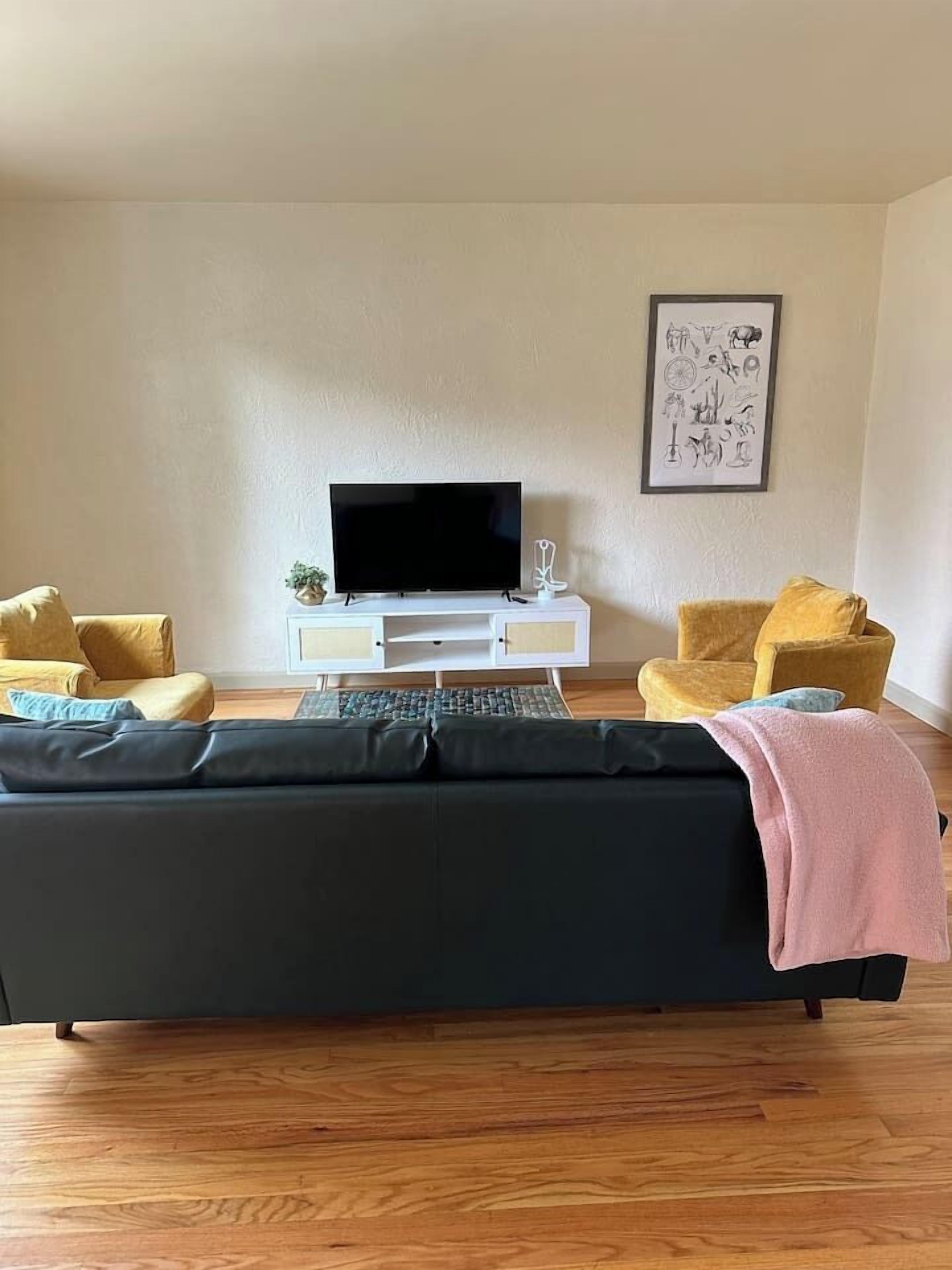 Een woonkamer met een zwarte bank, twee gele fauteuils, een witte tv-meubel en een ingelijst beeld aan de muur.