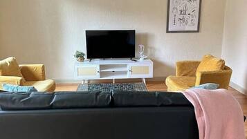 Een woonkamer met een zwarte bank, twee gele fauteuils, een witte tv-meubel en een ingelijst beeld aan de muur.