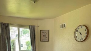 A dining area with a round table, four chairs, a window with curtains, a wall clock, and a framed picture.
