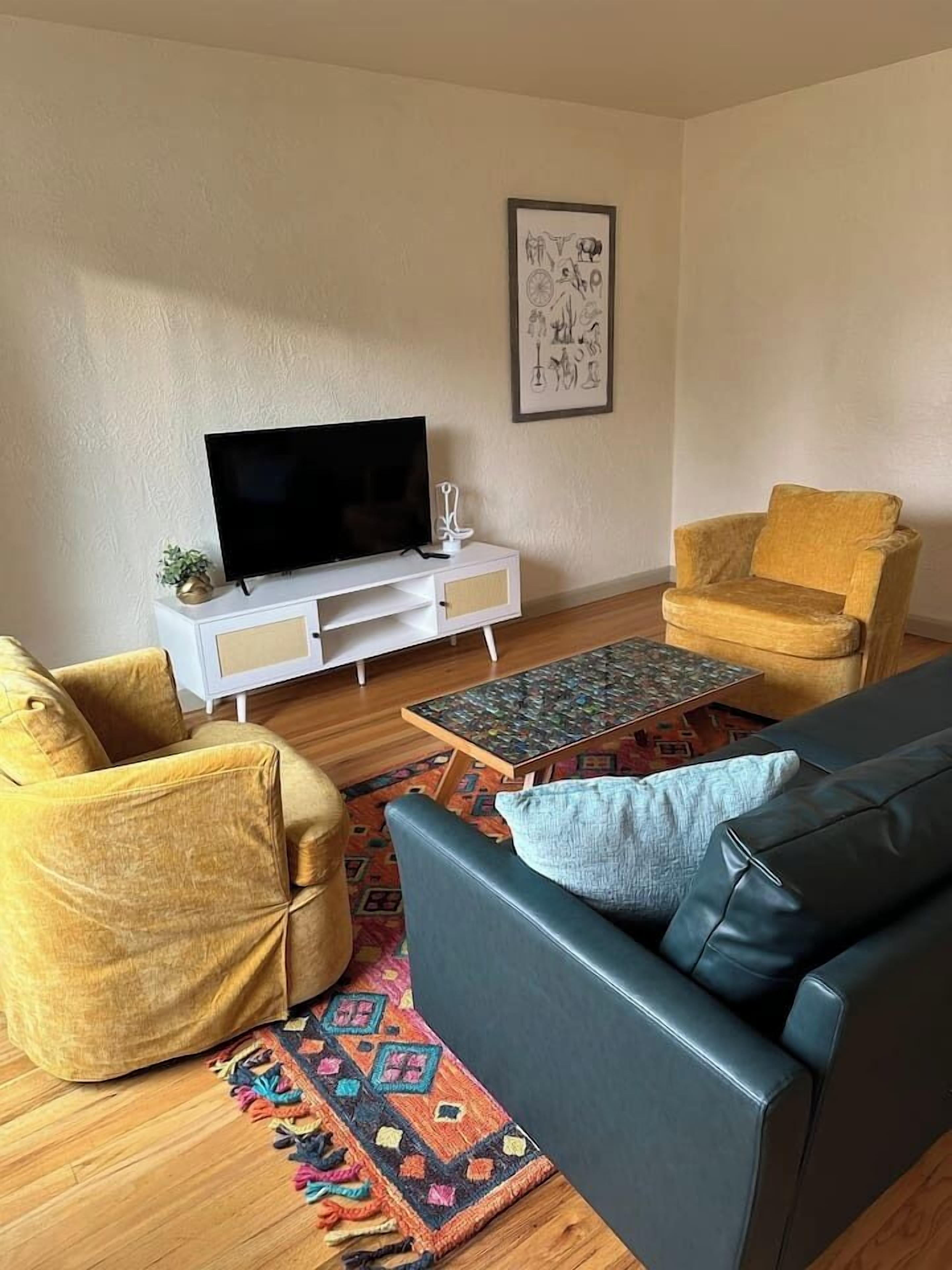 Un salon comprenant un canapé, deux fauteuils, une table basse, un téléviseur posé sur un meuble blanc et un tapis à motifs.