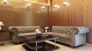 A modern lobby with two grey tufted sofas, a glass coffee table, and a wooden panel wall with recessed lighting. - Hotel Priydarshan (Kanpur)