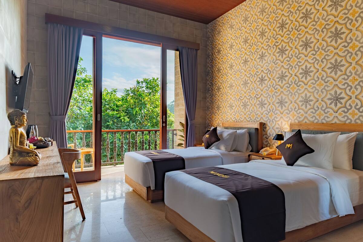 A hotel room with two beds, a desk, a Buddha statue, and a balcony with a view of greenery.