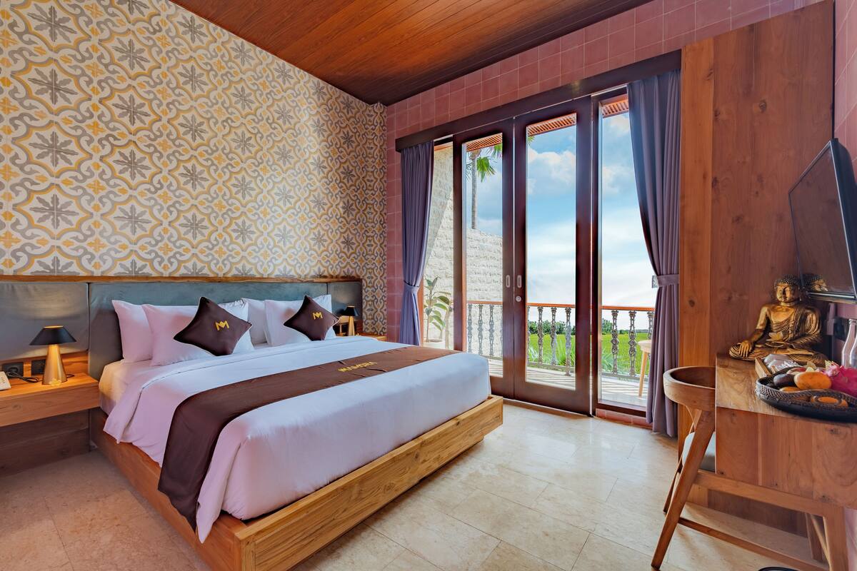 A spacious bedroom with a large bed, a wooden headboard, a nightstand with a lamp, and a view of the outdoors through a sliding glass door.