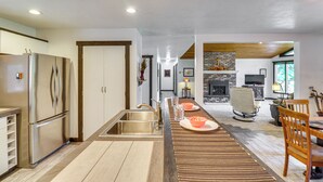A modern kitchen with stainless steel appliances, a wooden dining area with a long table, and a fireplace in the background. - Deck & Mtn Views: Libby Home on Golf Course! (Libby)