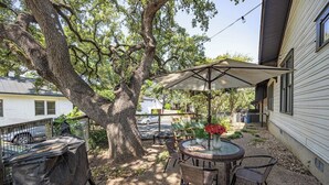 A backyard with a round table, chairs, and a large umbrella. There is a tree, a grill, and a house in the background.