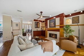 Ein Wohnzimmer mit Sofa, Sessel, Kamin, Deckenventilator und einem Essbereich im Hintergrund.
