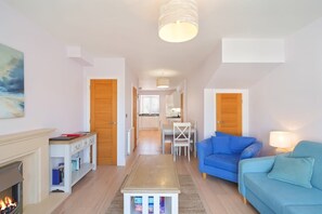A modern living room with a fireplace, a dining area with a table and chairs, a kitchen in the background, and a hallway leading to another room.