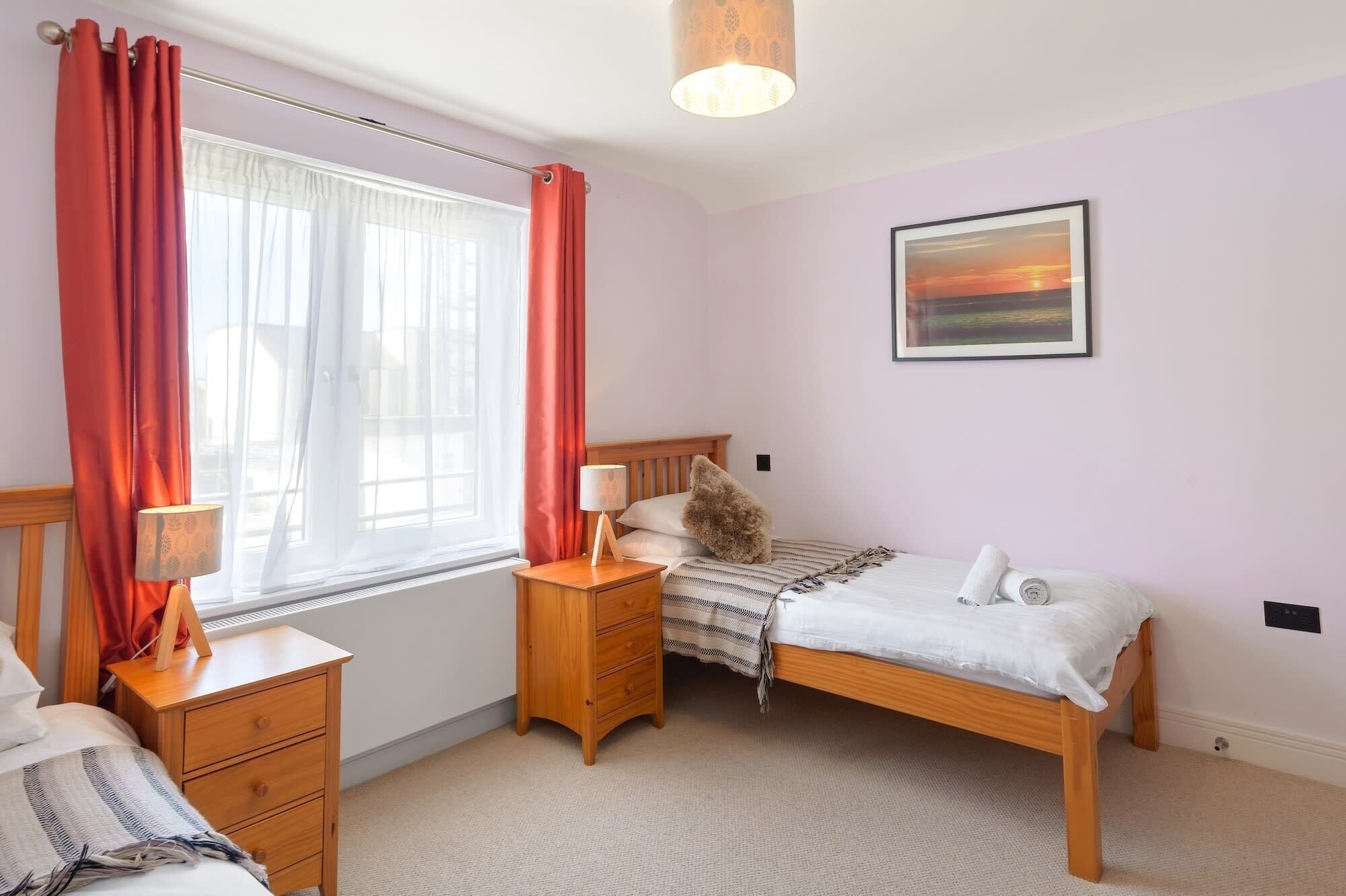Une chambre à coucher comprenant un lit, des tables de chevet, une lampe, une fenêtre avec des rideaux, un radiateur, un tableau accroché au mur et un sol recouvert d’une moquette.