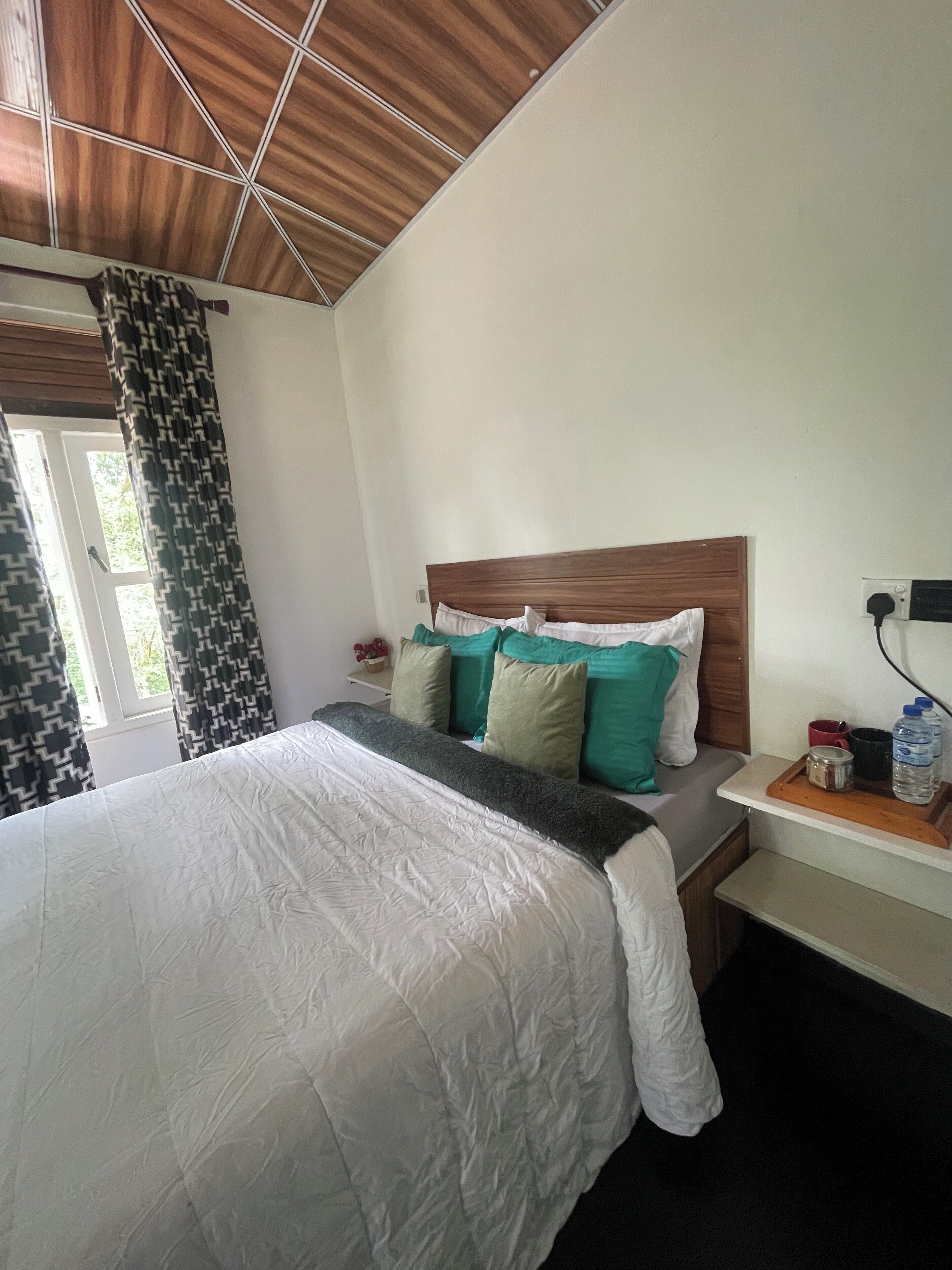 A neatly made bed with a wooden headboard, a bedside table with a water bottle and a red mug, and a window with patterned curtains.