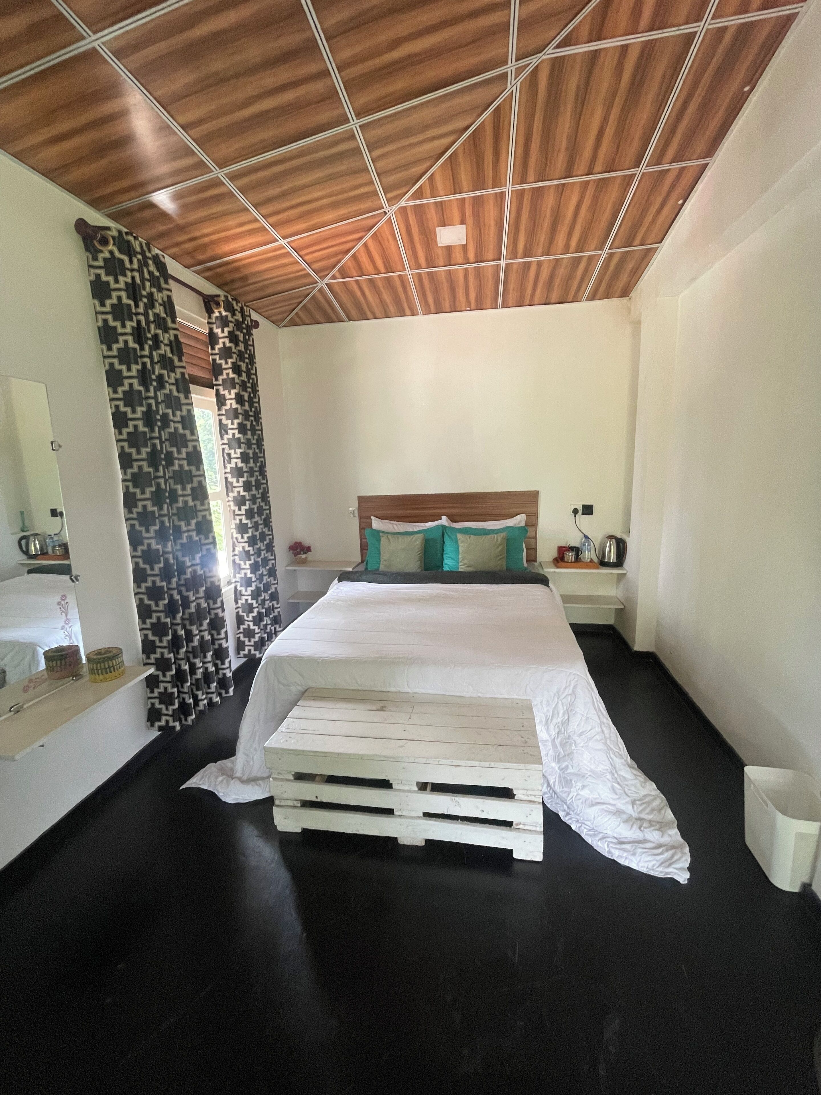 Un dormitorio con techo de madera, cama con colchón blanco y almohadas turquesa, suelo negro y pared blanca.