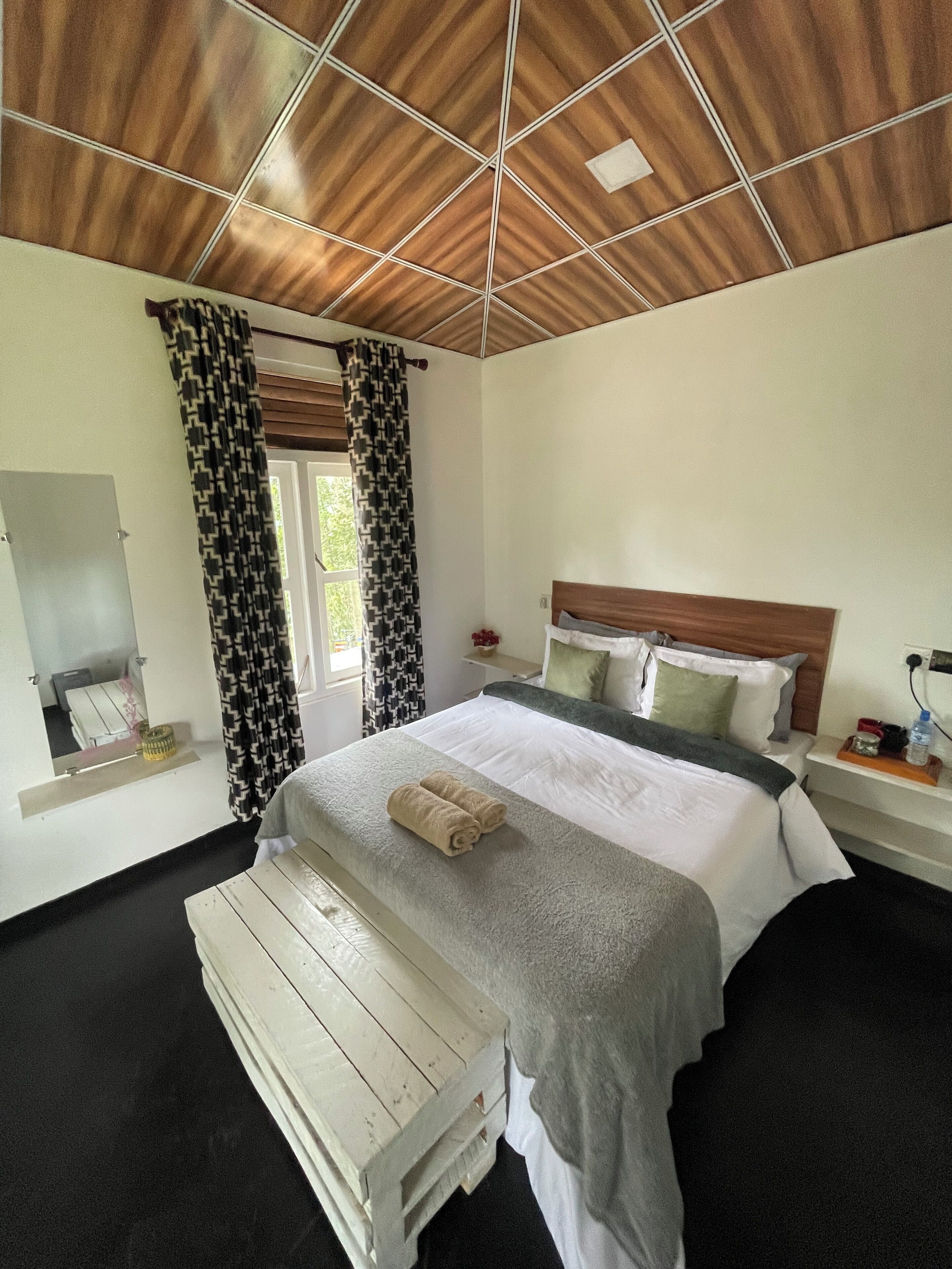 Une chambre avec un plafond en bois, un lit recouvert d’une couette grise et blanche, une table de chevet avec une lampe et une fenêtre ornée de rideaux à motifs.