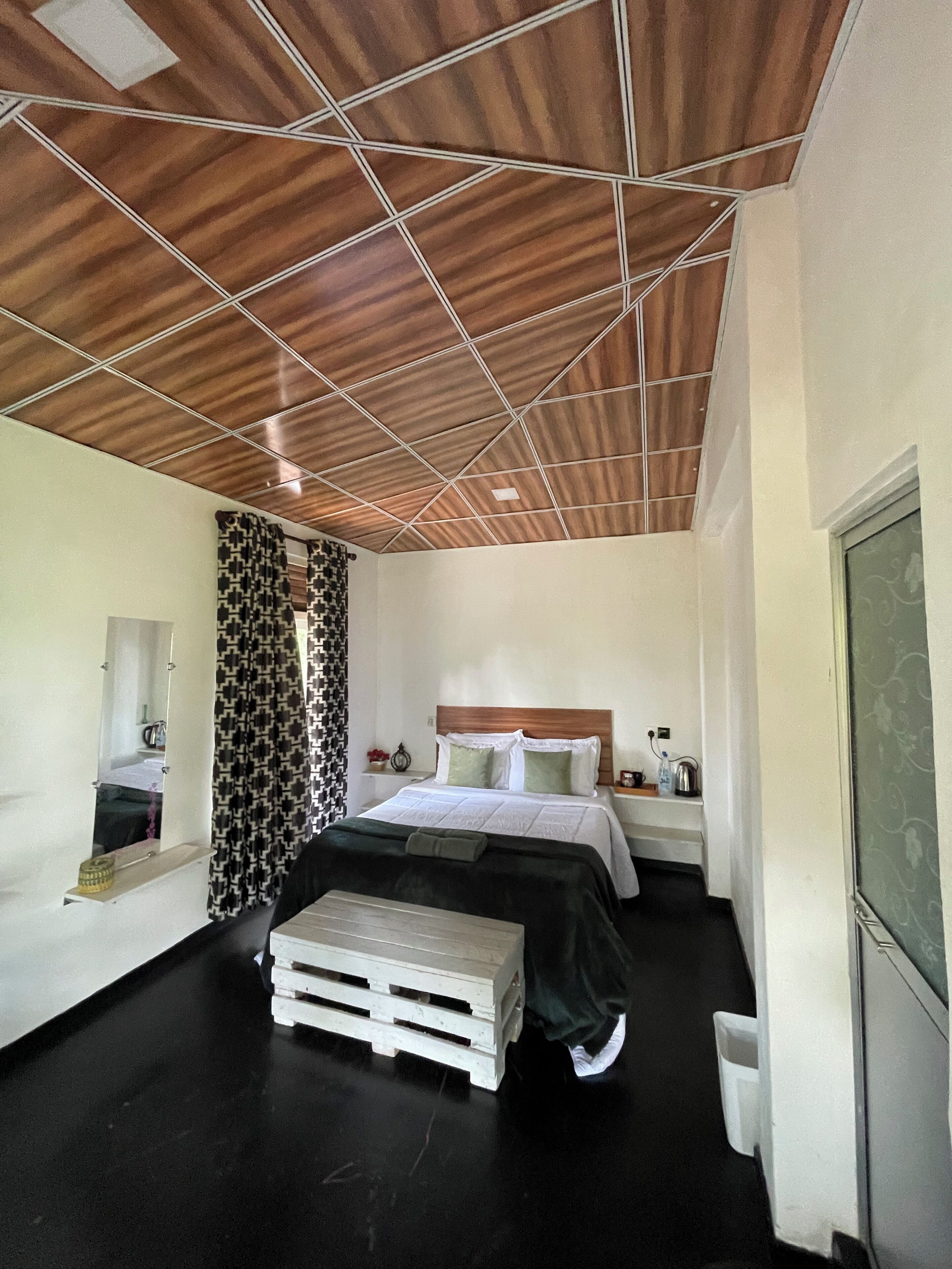 A hotel room with a wooden ceiling, a bed with white bedding, a bedside table with a lamp, and a window with a view.