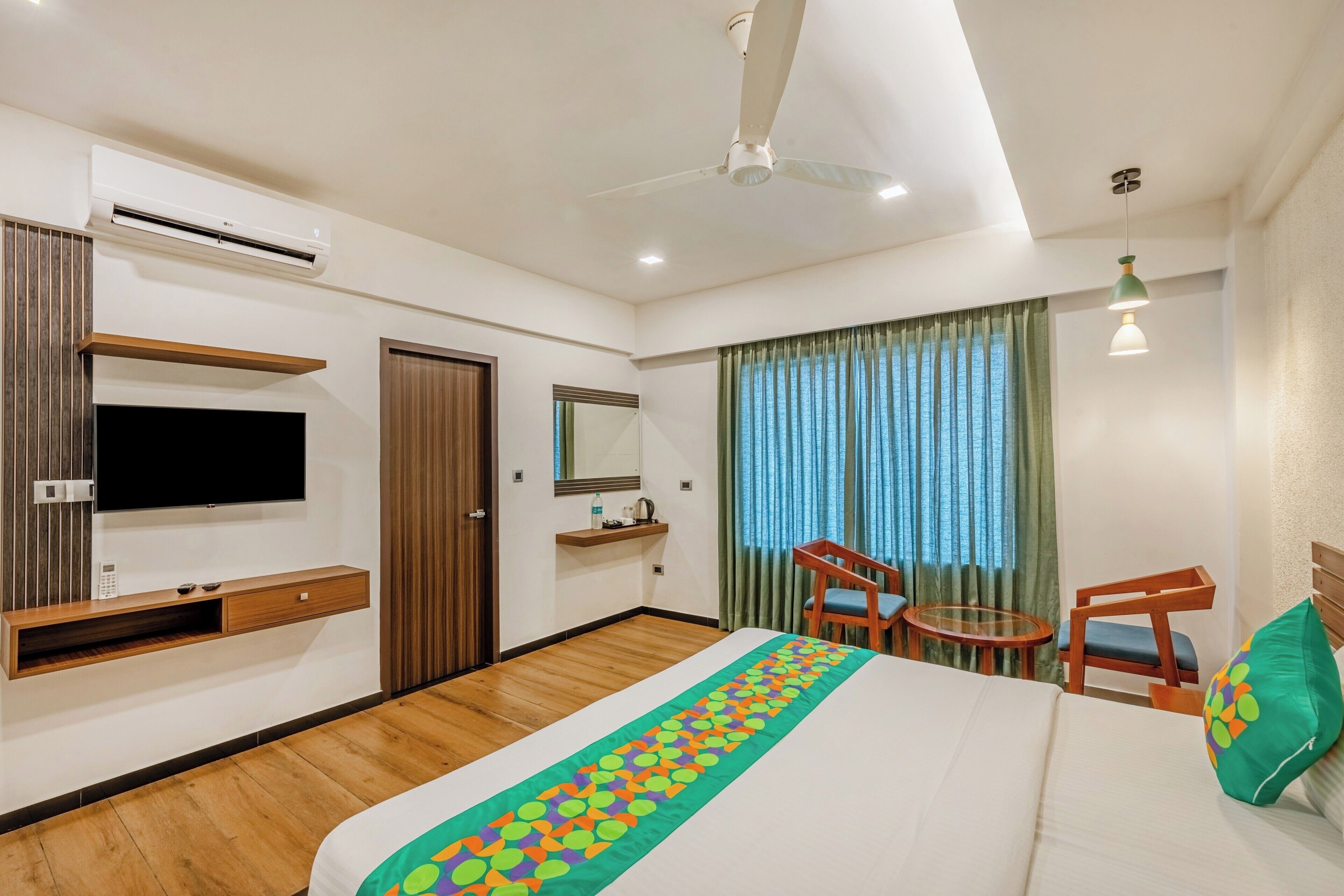 A hotel room with a bed, a TV, a desk, a chair, a small table, and a ceiling fan.