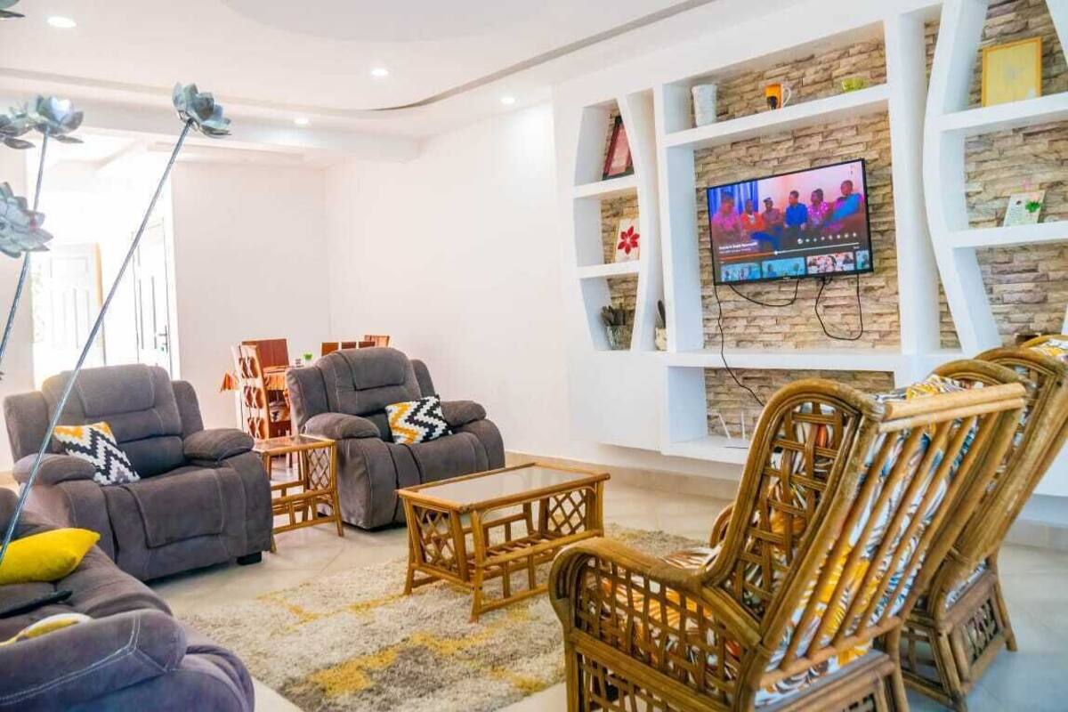 Un salon moderne avec un téléviseur à écran plat fixé à un mur en pierre, des canapés gris, une table basse et des fauteuils en osier.