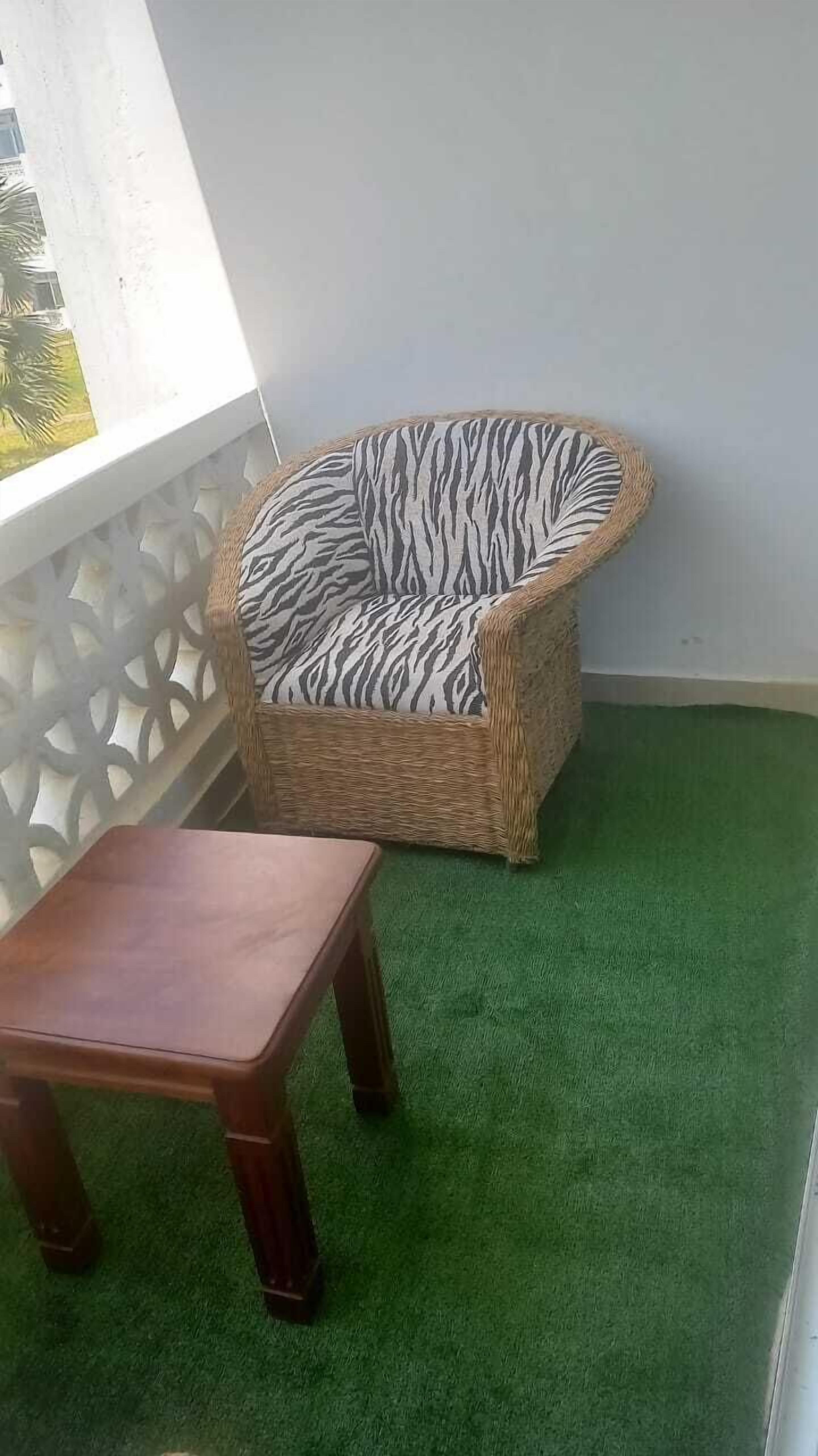 A wicker chair with a zebra print cushion, a wooden table, and a green artificial grass floor.