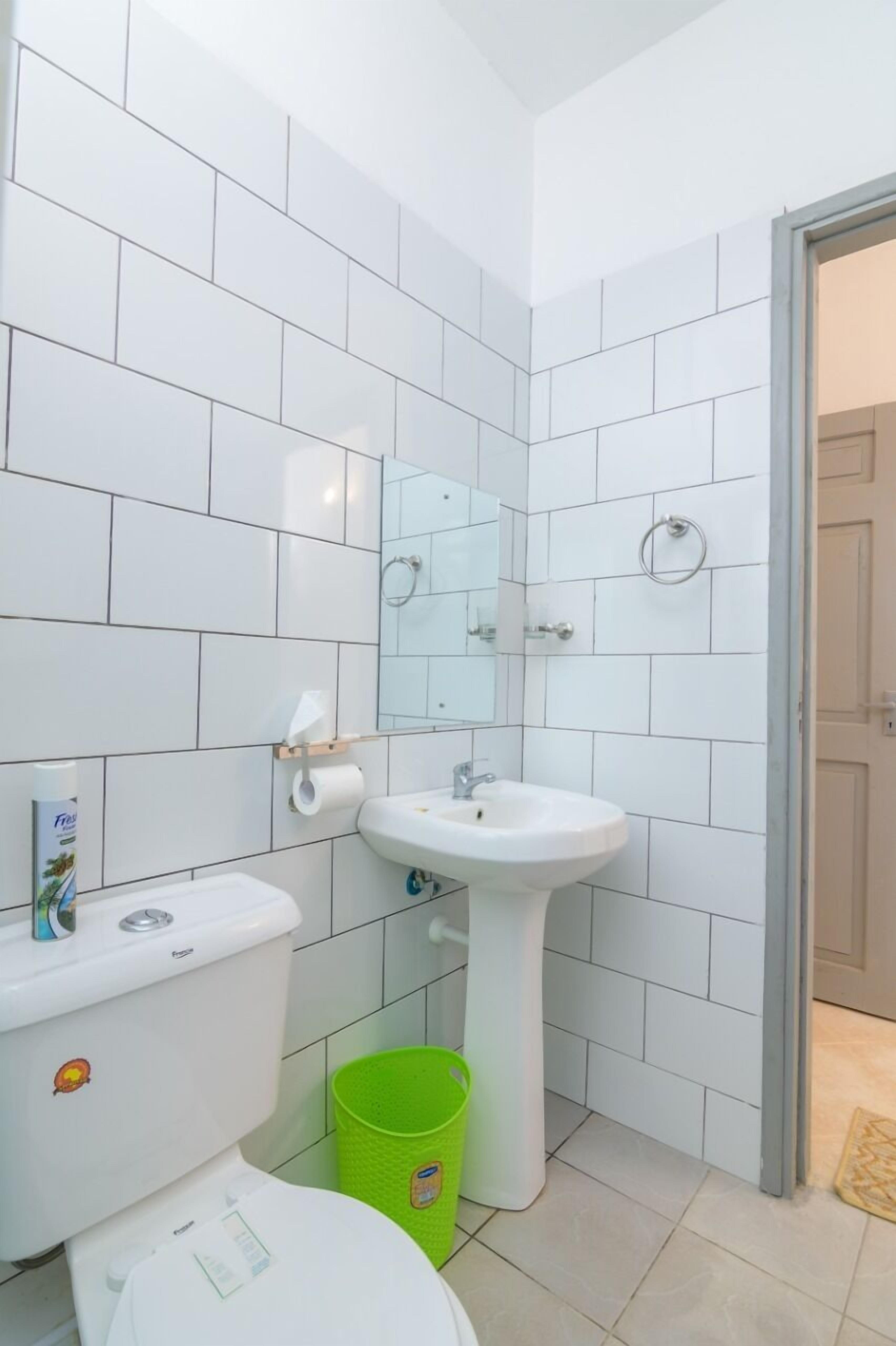 A bathroom with white tiles, a white sink, a toilet, and a green trash bin.
