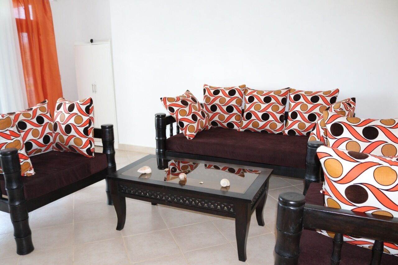 Uma sala de estar com um sofá e duas poltronas, cada uma com almofadas estampadas, uma mesa de centro de vidro com objetos decorativos, e uma porta branca ao fundo.