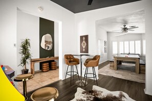 A modern living room with a round table, two bar stools, a ceiling fan, and a large window.
