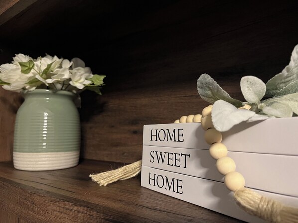 A wooden shelf with a white beaded string, a green vase with white flowers, and a white box with the words "HOME SWEET HOME" on it.
