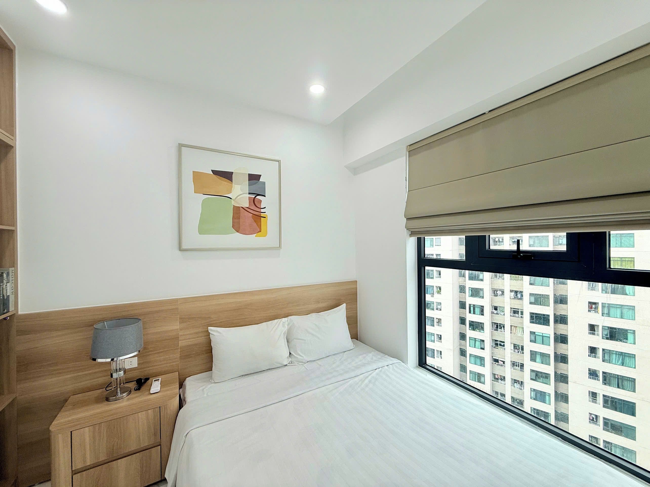 A modern hotel room with a bed, bedside table, lamp, and a window with a city view.