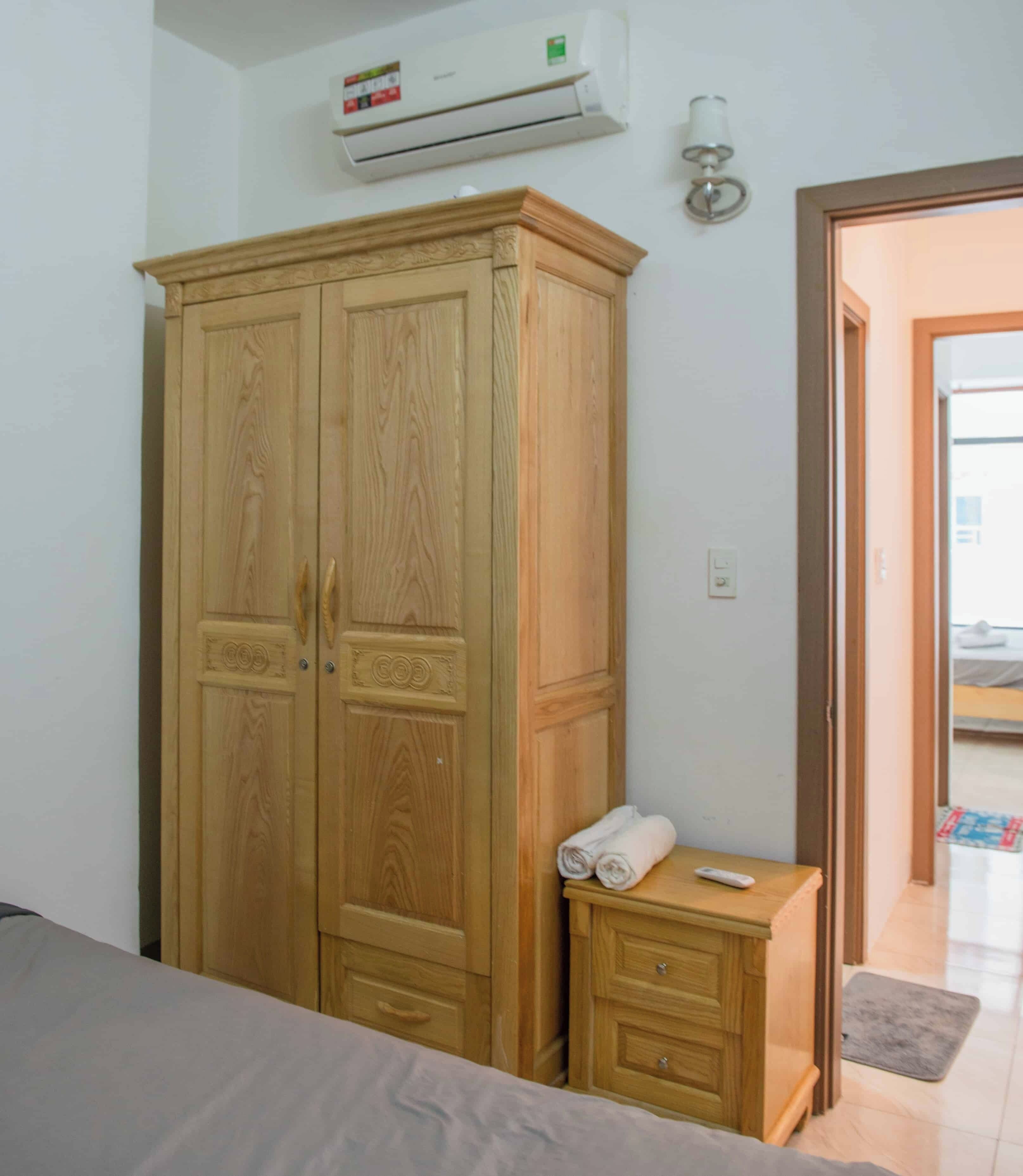 A bedroom with a wooden wardrobe, a bedside table with towels, and an air conditioning unit on the wall.