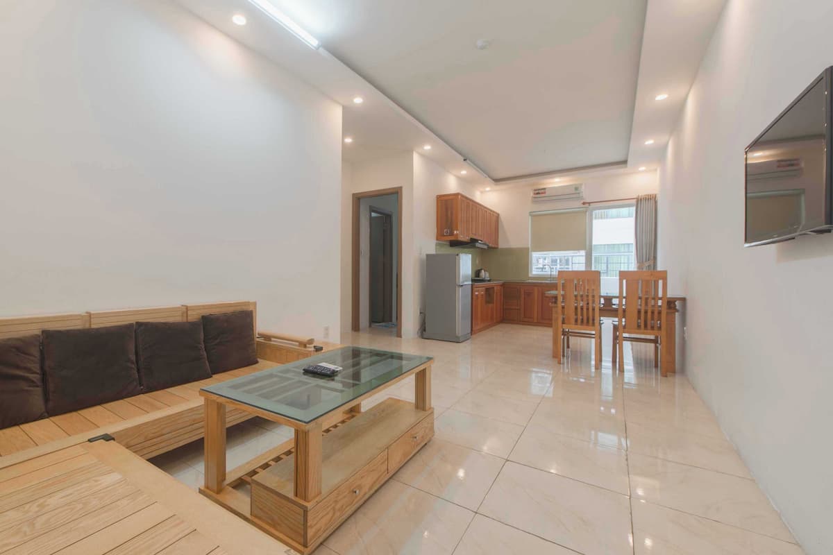 A modern living room with a wooden sofa, glass coffee table, and wooden chairs. There is a kitchen area with wooden cabinets and a refrigerator.