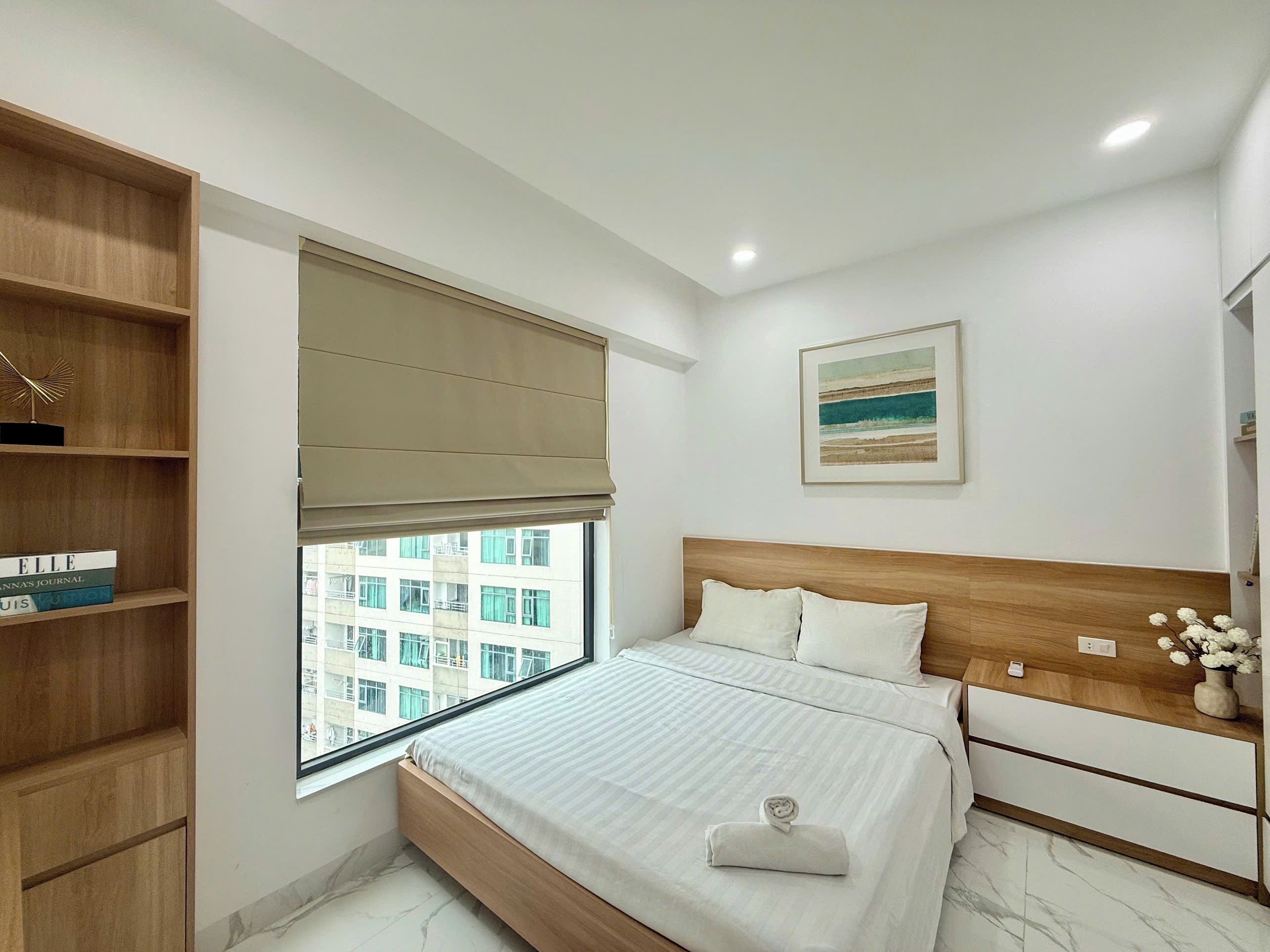A bedroom with a bed, a bedside table, a window with a view of buildings, a bookshelf, and a painting on the wall.