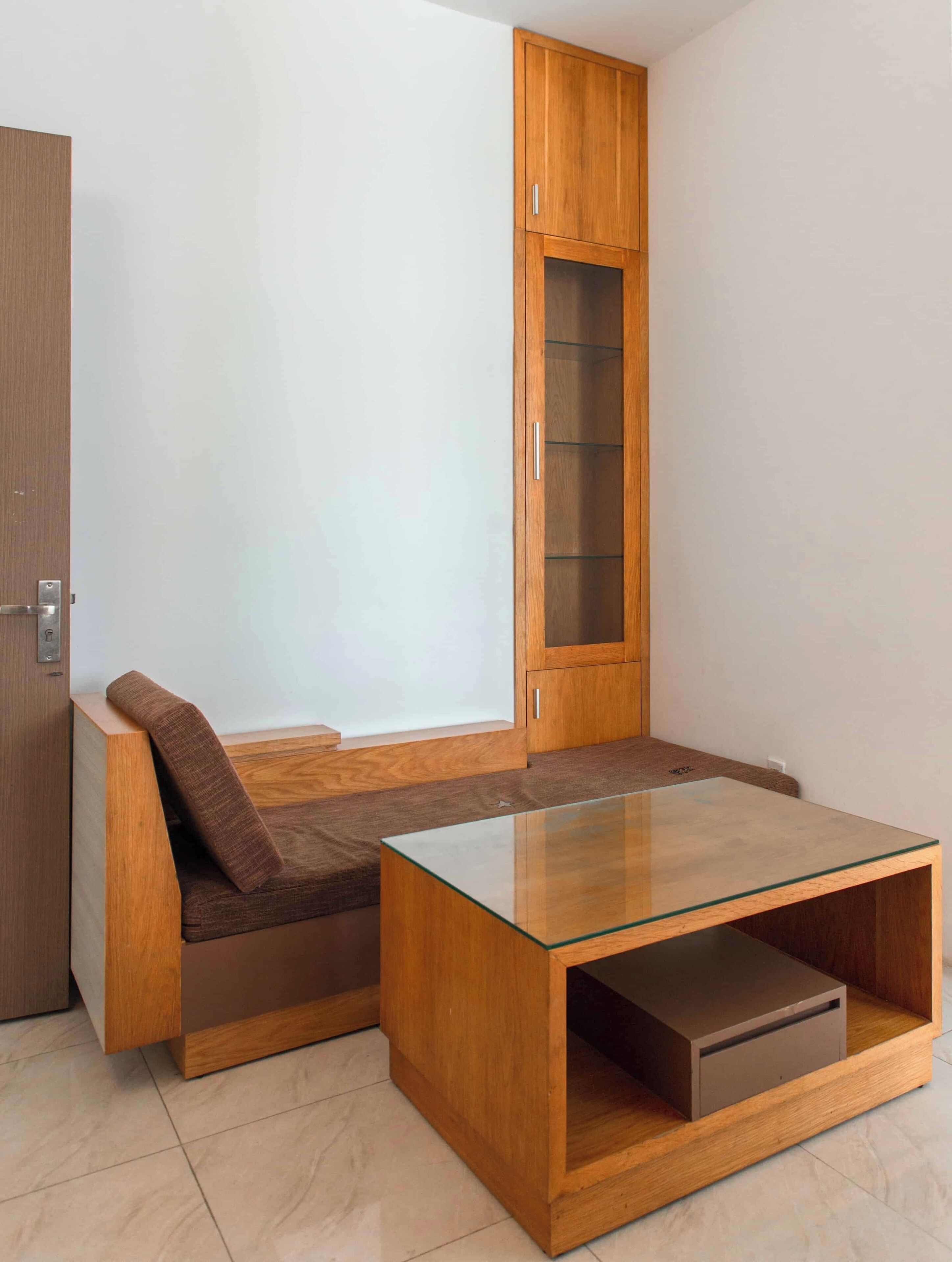 A modern room with a built-in bench seating area, a glass-top coffee table, and a wooden cabinet with a glass door.