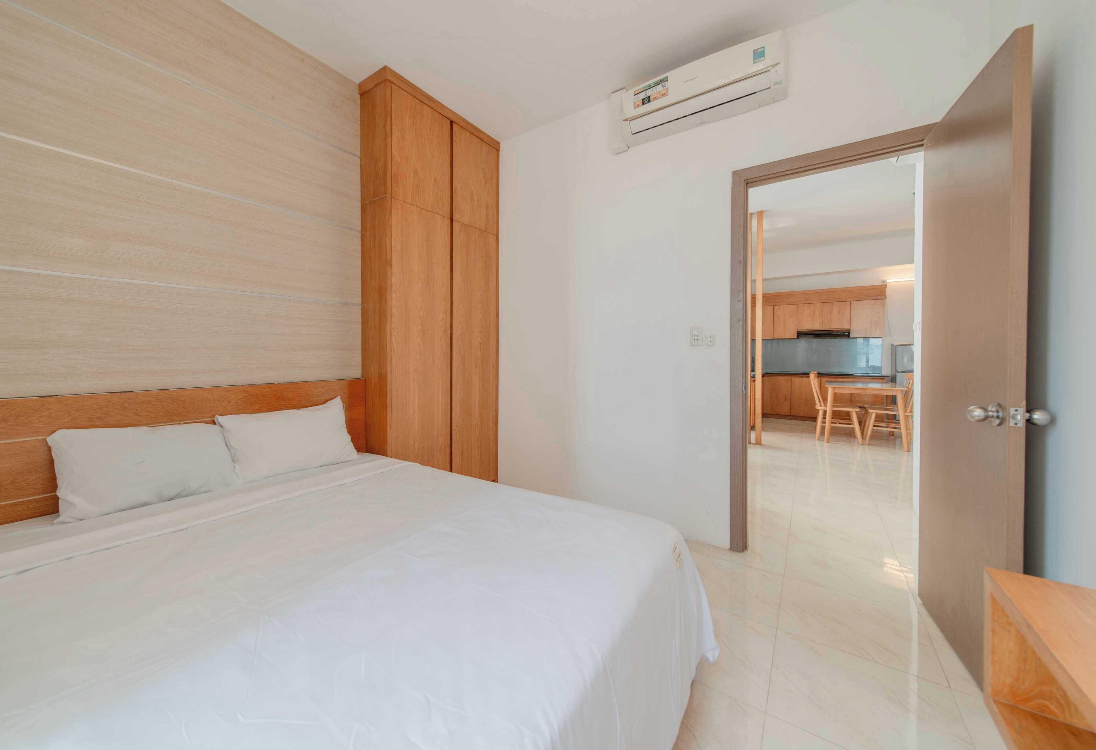 A hotel room with a wooden headboard, a bed with white linens, a wooden wardrobe, and a kitchenette visible through an open door.