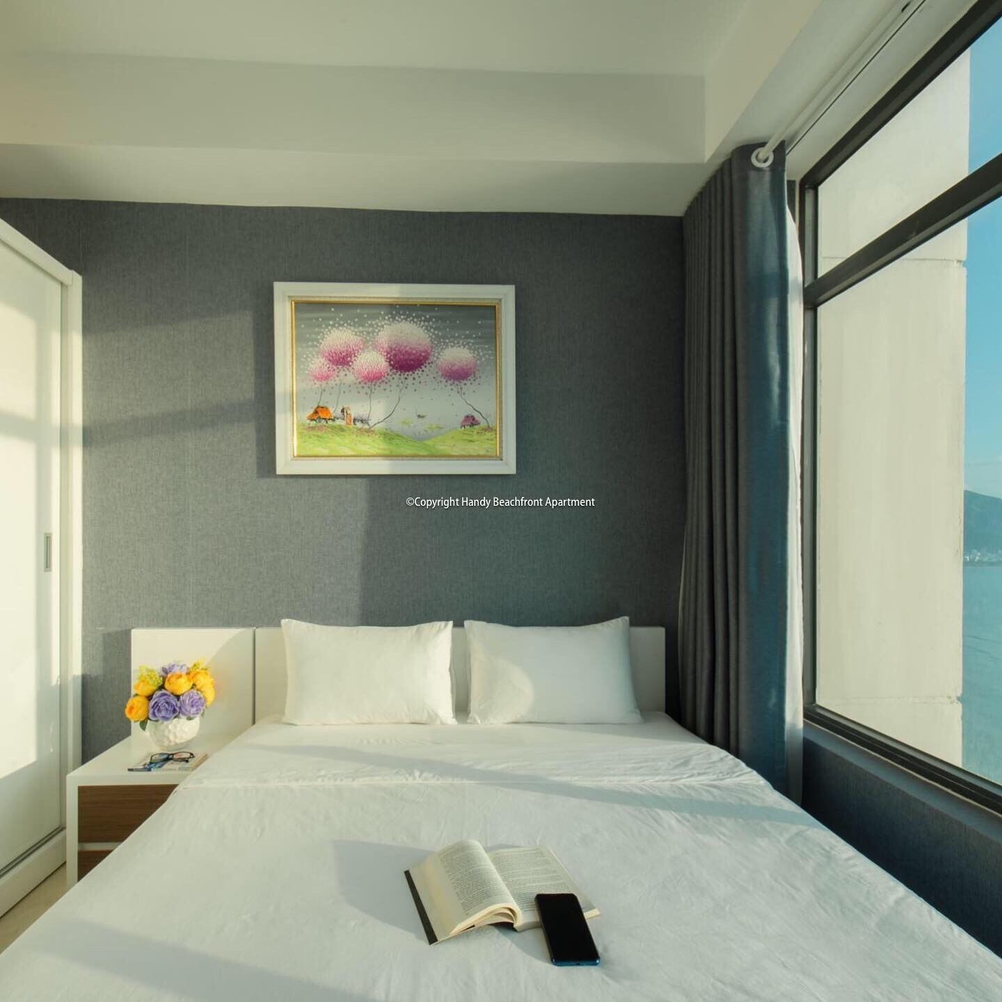 A hotel room with a bed, a nightstand with a vase of flowers, a framed picture on the wall, and a large window.