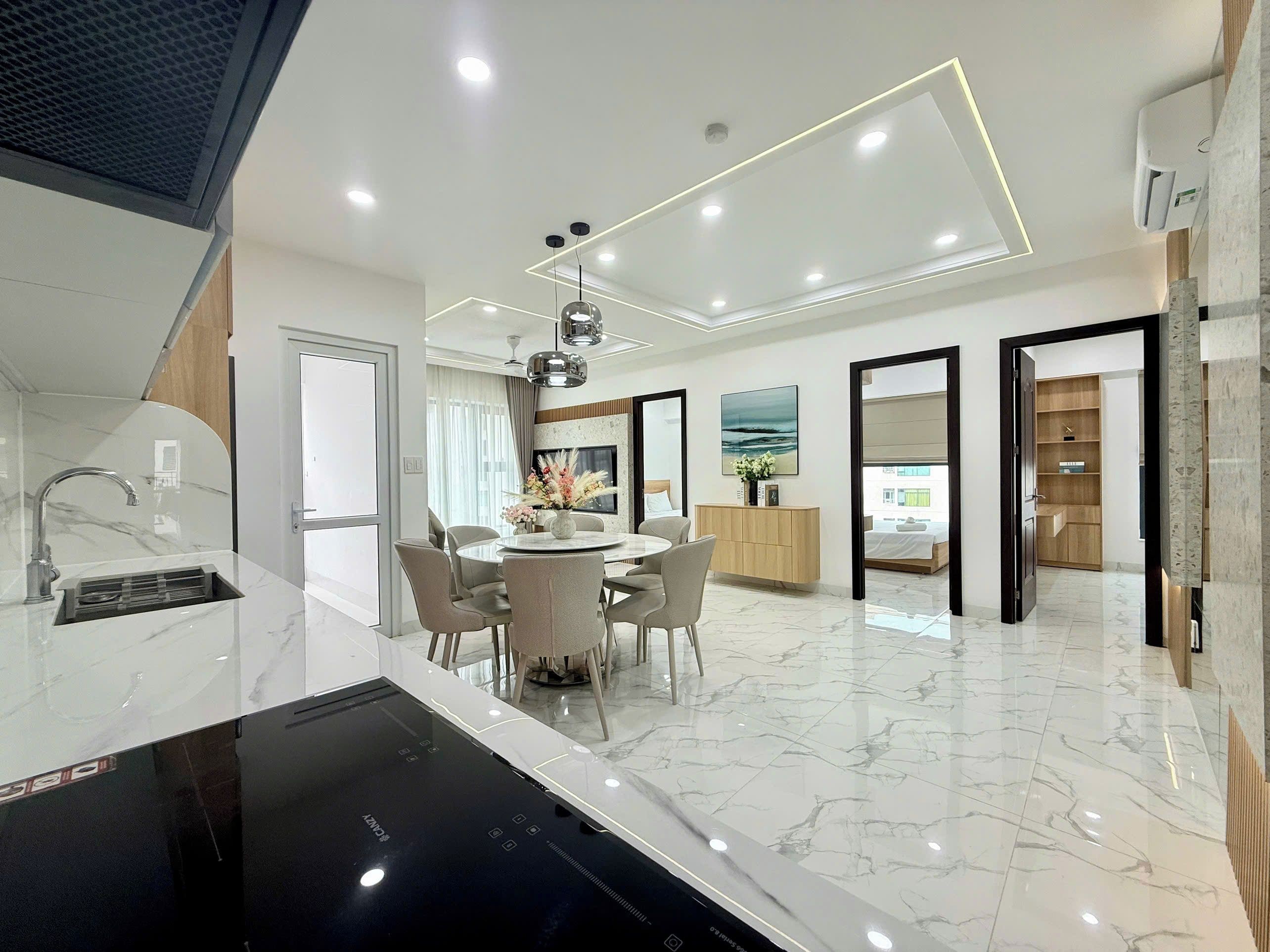 A modern kitchen with a built-in stove, a dining area with a round table and chairs, and a living space with a TV and wooden shelving.