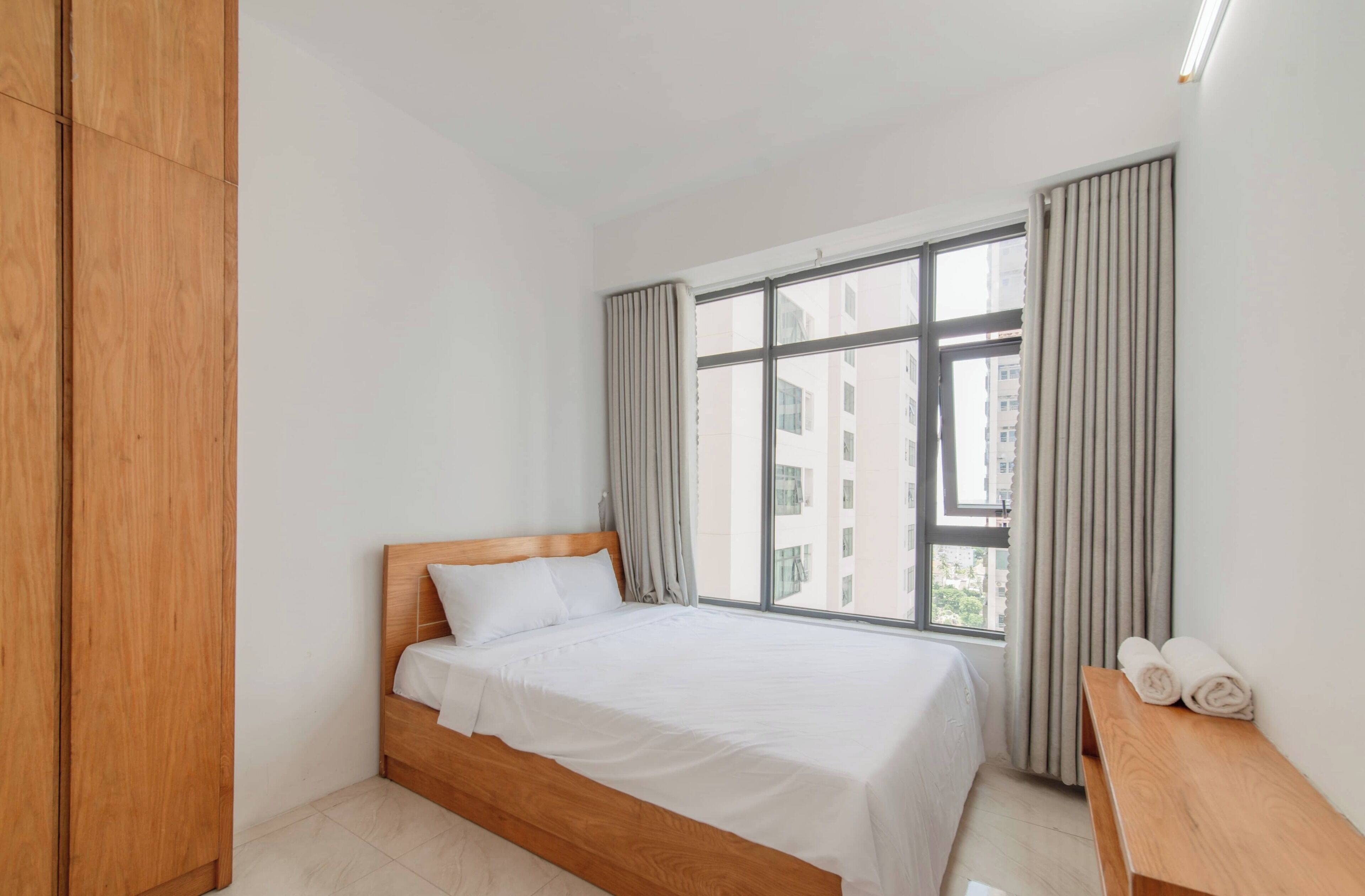 A modern hotel room with a wooden bed, a large window with curtains, and a wooden bench with towels.