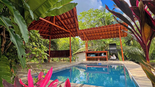 A pool area with a wooden deck, a covered seating area, and lush tropical plants. - La Casa de Piedra: Un Hogar Único (Puerto Jiménez)