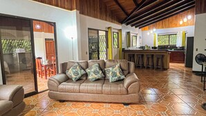 A spacious living area with a wooden ceiling, a brown leather sofa with patterned cushions, a coffee table with a bowl and remote, and a kitchen in the background. - La Casa de Piedra: Un Hogar Único (Puerto Jiménez)