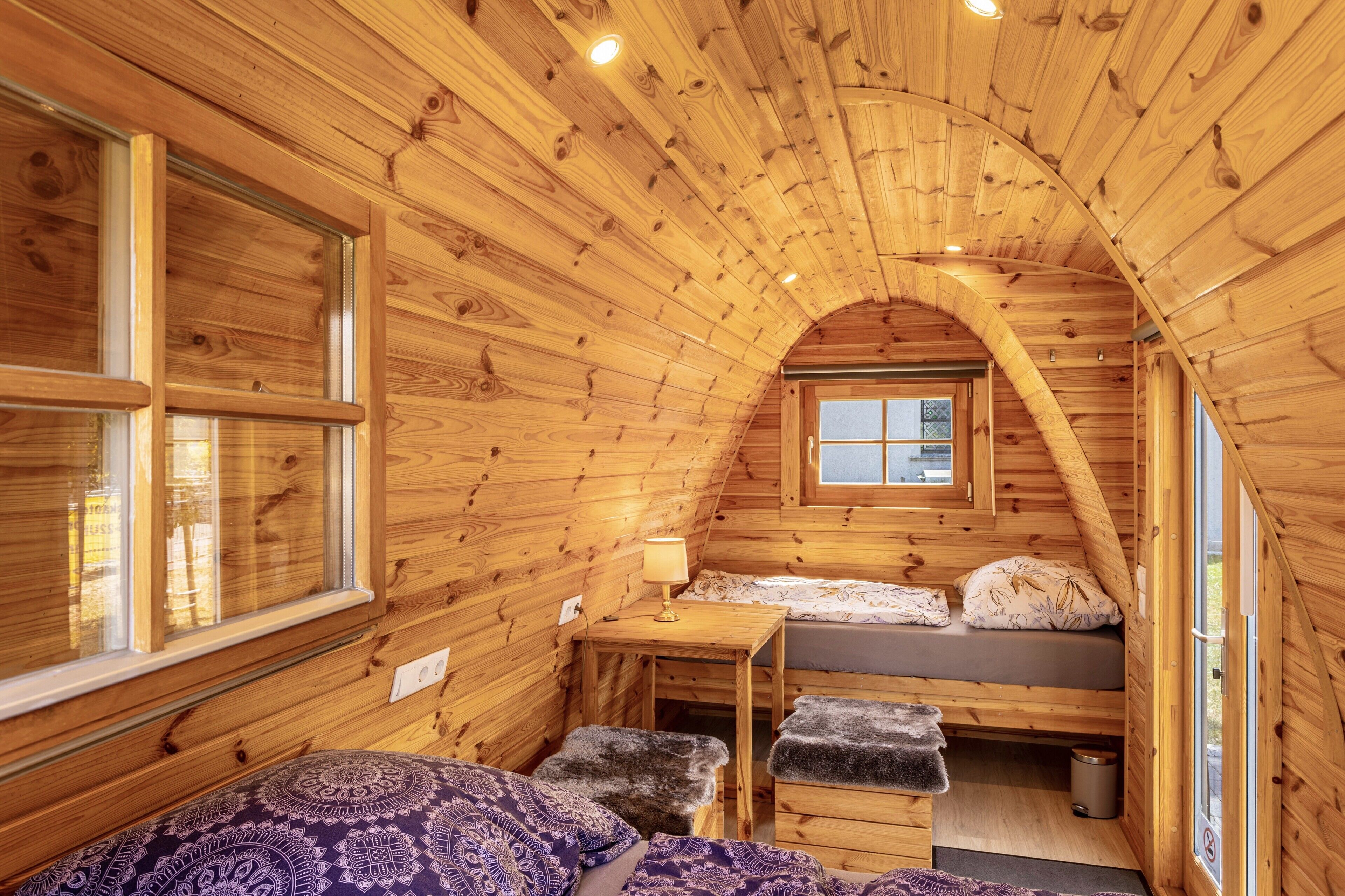Une cabane en bois avec une chambre à coucher située en mezzanine, une petite table avec une lampe et une fenêtre.