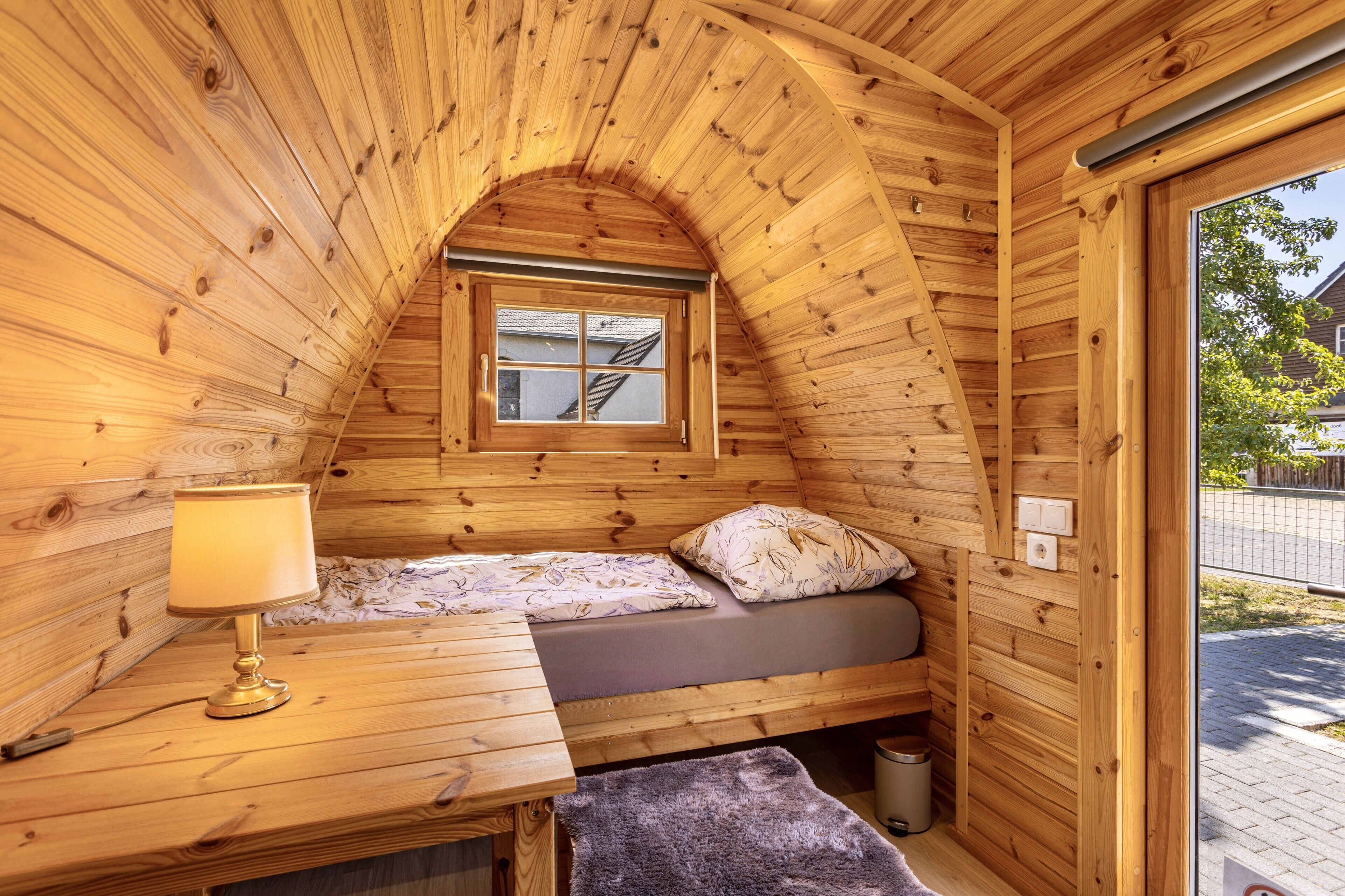 Um pequeno quarto de madeira com teto inclinado, uma cama simples com travesseiro e cobertor, uma mesa de cabeceira com abajur, e uma janela com vista para o exterior.